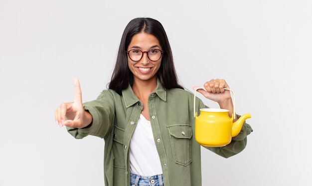 Junge hispanische Frau, die stolz und selbstbewusst lächelt, die Nummer eins macht und eine Teekanne hält