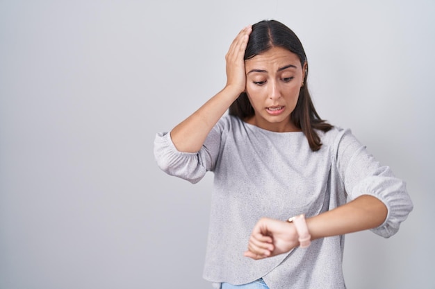 Junge hispanische Frau, die über weißem Hintergrund steht und auf die Uhrzeit schaut, besorgt, Angst, zu spät zu kommen