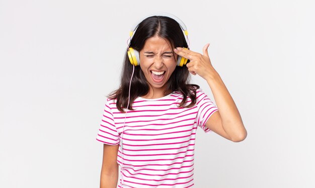 Junge hispanische Frau, die unglücklich und gestresst aussieht, Selbstmordgeste, die Waffenzeichen mit Kopfhörern hört