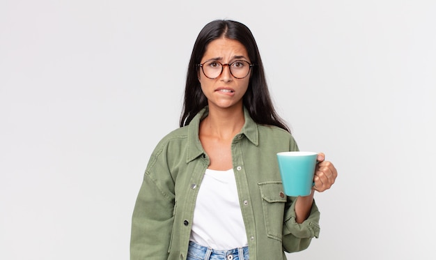 Junge hispanische Frau, die verwirrt und verwirrt aussieht und eine Kaffeetasse hält