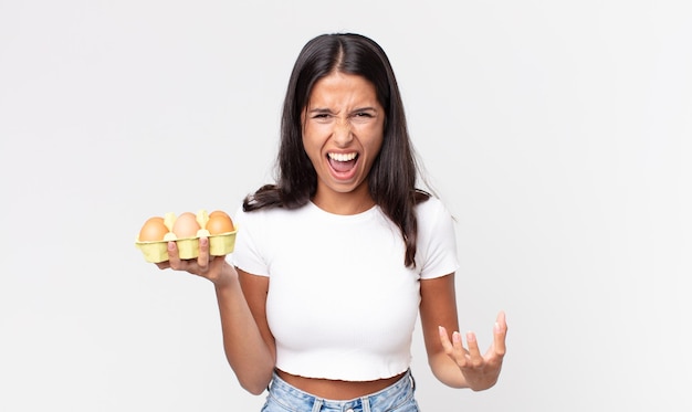 Junge hispanische Frau, die wütend, verärgert und frustriert aussieht und eine Eierschachtel hält