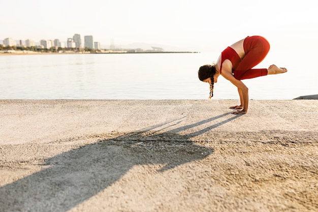 Junge hispanische Frau, die Yoga praktiziert und Bakasana-Position am Meer macht - Crane Pose Yoga am Strand morgens - Wellness- und Gesundheitskonzept