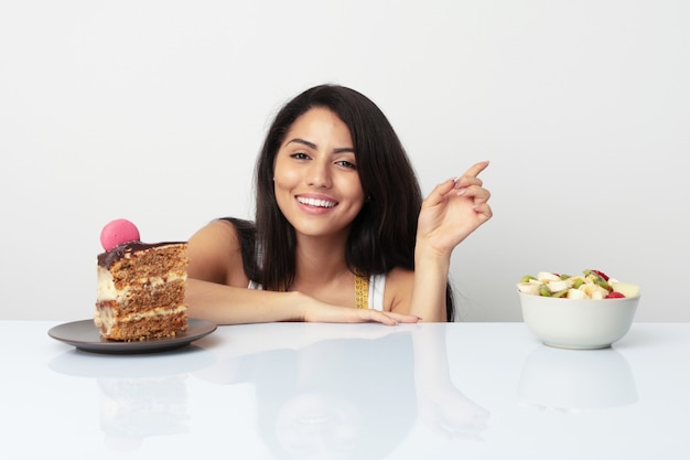 Junge hispanische Frau, die zwischen dem Kuchen- oder Fruchtlächeln freundlich zeigt mit dem Zeigefinger weg wählt.