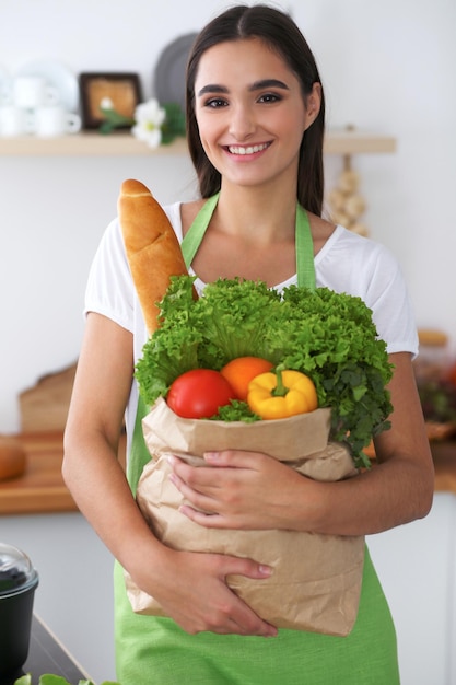 Junge hispanische Frau in einer grünen Schürze, die in der Küche kocht Hausfrau, die eine Papiertüte voller Gemüse und Obst hält, während sie lächelt Gesunde Ernährung und vegetarisches Konzept