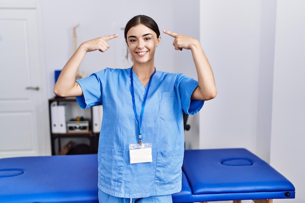 Junge hispanische Frau in Physiotherapeutenuniform, die lächelnd in der Klinik steht und mit beiden Händen auf den Kopf zeigt, eine tolle Idee oder ein gutes Gedächtnis