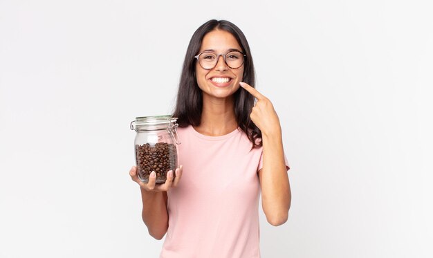 Junge hispanische Frau lächelt selbstbewusst, zeigt auf ein breites Lächeln und hält eine Kaffeebohnenflasche