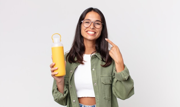 Junge hispanische Frau lächelt selbstbewusst, zeigt auf ein breites Lächeln und hält eine Kaffeethermoskanne