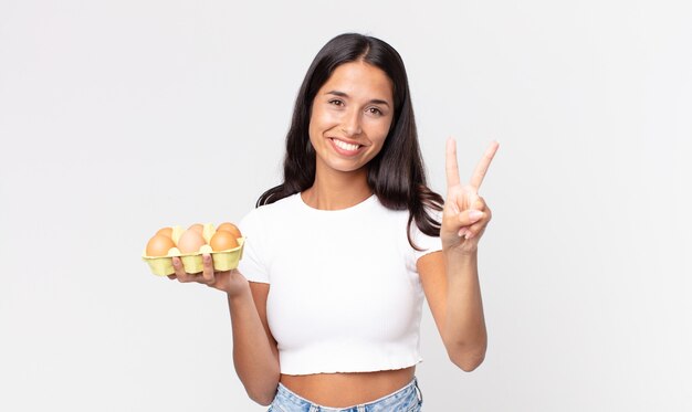 Junge hispanische Frau lächelt und sieht freundlich aus, zeigt Nummer zwei und hält eine Eierschachtel