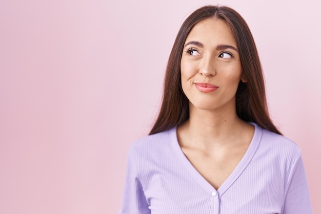 Junge hispanische Frau mit langen Haaren, die über rosafarbenem Hintergrund steht, lächelnd zur Seite schaut und nachdenklich wegstarrt