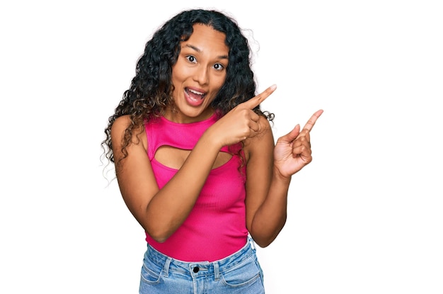 Junge hispanische Frau mit lockigem Haar, rosafarbenem Oberteil, lächelnd in die Kamera schauend, mit zwei Händen und Fingern zur Seite zeigend