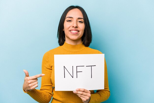 Junge hispanische Frau mit NFT-Plakat isoliert auf blauem Hintergrund, die mit der Hand auf einen Hemdkopienraum zeigt, stolz und selbstbewusst