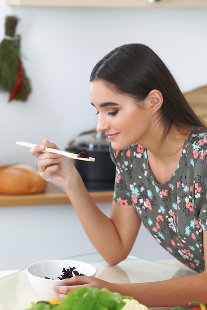 Junge hispanische Frau oder Studentin, die in der Küche kocht Mädchen schmeckt frischen Salat, während sie am Tisch sitzt