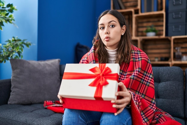 Junge hispanische Frau öffnet die Geschenkbox und macht das Fischgesicht mit Mund und zusammengekniffenen Augen verrückt und komisch