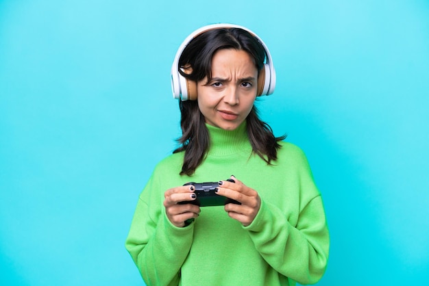 Foto junge hispanische frau spielt mit einem videospiel-controller, isoliert auf blauem hintergrund und hat zweifel