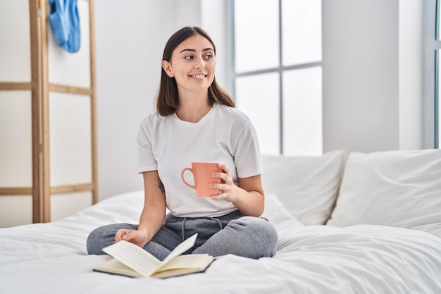 Junge hispanische Frau trinkt eine Tasse Kaffee und liest ein Buch im Schlafzimmer