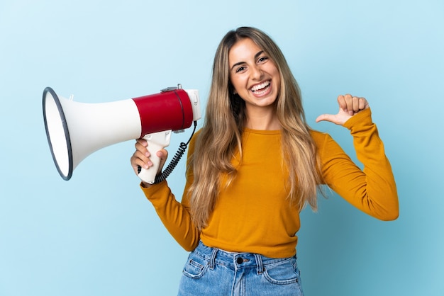 Junge hispanische Frau über isoliertem Blau, das ein Megaphon hält und stolz und selbstzufrieden ist