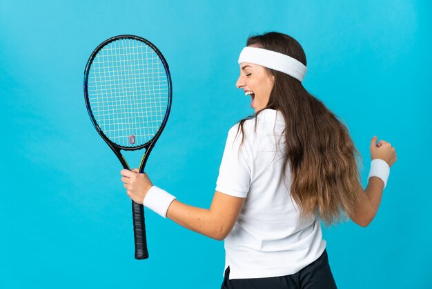 Junge hispanische Frau über isoliertem blauem Hintergrund, die Tennis spielt und einen Sieg feiert