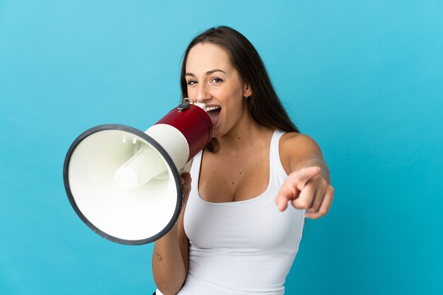 Junge hispanische Frau über isolierter blauer Wand, die durch ein Megaphon schreit, um etwas anzukündigen, während sie nach vorne zeigt