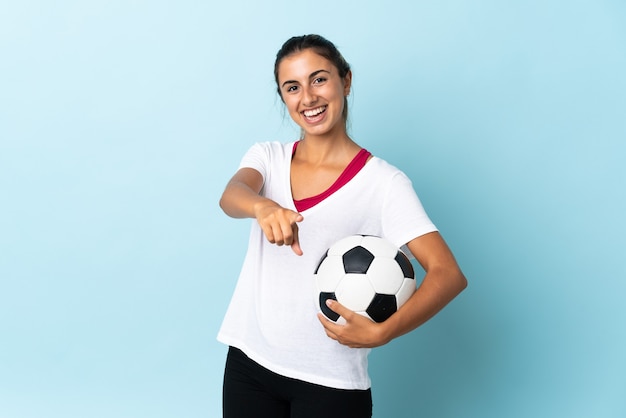 Junge hispanische Frau über isolierter blauer Wand mit Fußball und nach vorne zeigend