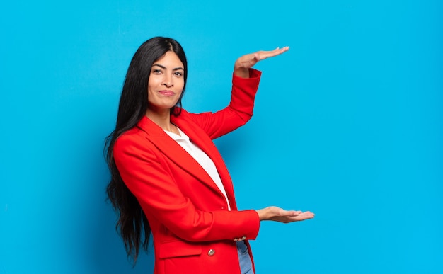 Junge hispanische Geschäftsfrau, die ein Objekt mit beiden Händen auf seitlichem Kopierraum hält, ein Objekt zeigt, anbietet oder bewirbt
