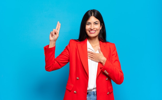 Junge hispanische Geschäftsfrau, die glücklich, selbstbewusst und vertrauenswürdig aussieht, lächelt und ein Siegeszeichen zeigt, mit einer positiven Einstellung