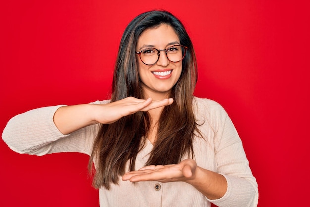 Junge hispanische, kluge Frau mit Brille, die vor rotem, isoliertem Hintergrund steht und mit Händen gestikuliert, die große und große Maßzeichen zeigen, lächelt lächelnd in die Kamera und misst Konzept