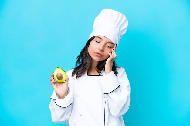 Junge hispanische Köchin, die Avocado isoliert auf blauem Hintergrund mit Kopfschmerzen hält