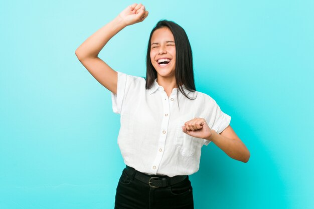 Junge hispanische kühle Frau gegen eine blaue Wand, die einen speziellen Tag feiert, springt und hebt Arme mit Energie an.