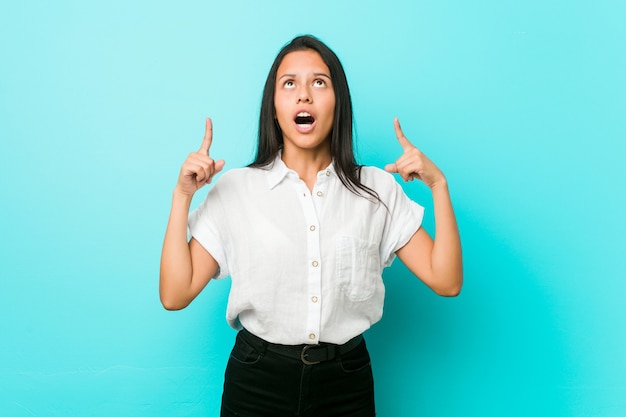 Junge hispanische kühle Frau gegen eine blaue Wand, die oben mit geöffnetem Mund zeigt.