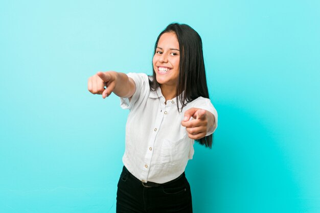 Junge hispanische kühle Frau gegen eine blaue Wand fröhliches Lächeln zeigt nach vorne.