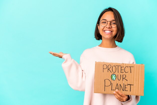 Junge hispanische Mischrassenfrau, die einen Schutzkarton unseres Planeten hält, der einen Kopienraum auf einer Handfläche zeigt und eine andere Hand auf Taille hält.