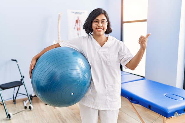 Junge hispanische Physiotherapeutin hält Pilates-Ball in der Hand und zeigt mit dem Daumen zur Seite, lächelt glücklich und öffnet den Mund