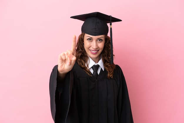 Junge Hochschulabsolventin isoliert auf rosa Hintergrund, die einen Finger zeigt und hebt