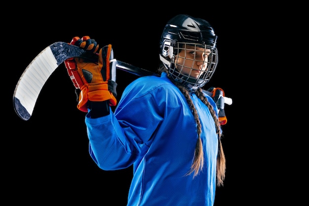 Junge Hockeyspielerin auf schwarzem Hintergrund isoliert. Tragende Ausrüstung und Helm der Sportlerin, die mit dem Stock stehen. Konzept des Sports, gesunder Lebensstil, Bewegung, Bewegung, Aktion.