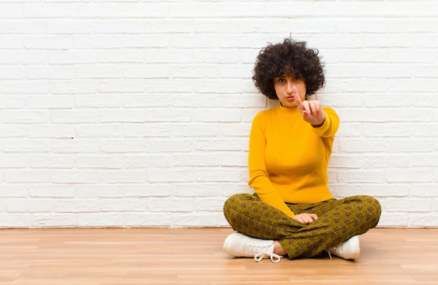 Junge hübsche Afro-Frau, die stolz und zuversichtlich lächelt und die Nummer eins triumphierend posiert und sich wie ein Anführer fühlt, der auf dem Boden sitzt