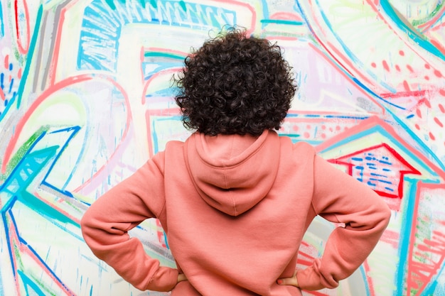 Junge hübsche Afrofrau, die verwirrt oder voll oder die Zweifel und Fragen sich fühlen und wundern, mit den Händen auf Hüften, hintere Ansicht gegen Graffitiwand