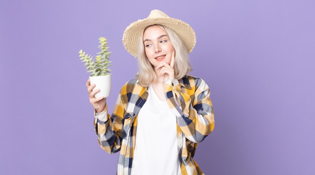 Junge hübsche Albino-Frau, die glücklich lächelt und träumt oder zweifelt und einen Zimmerpflanzenkaktus hält