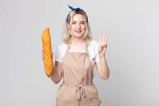 Junge hübsche Albino-Frau, die lächelt und freundlich aussieht und Nummer drei mit einem Brotbaguette zeigt
