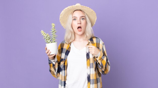 Junge hübsche Albino-Frau, die schockiert und überrascht mit weit geöffnetem Mund aussieht, auf sich selbst zeigt und einen Zimmerpflanzenkaktus hält