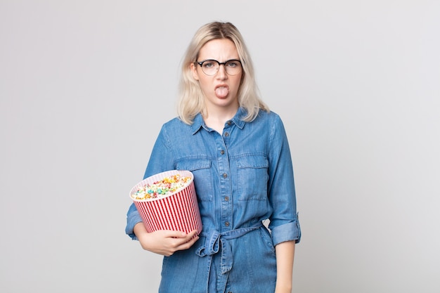 Junge hübsche Albino-Frau, die sich angewidert und gereizt fühlt und die Zunge mit einem Popcorn-Eimer herausstreckt?