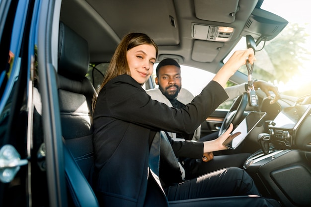 Junge hübsche Autohändlerin der kaukasischen Frau, die Autoschlüssel und digitales Tablett hält, während sie im Auto mit ihrem lächelnden hübschen Kunden, afrikanischen Geschäftsmann sitzt