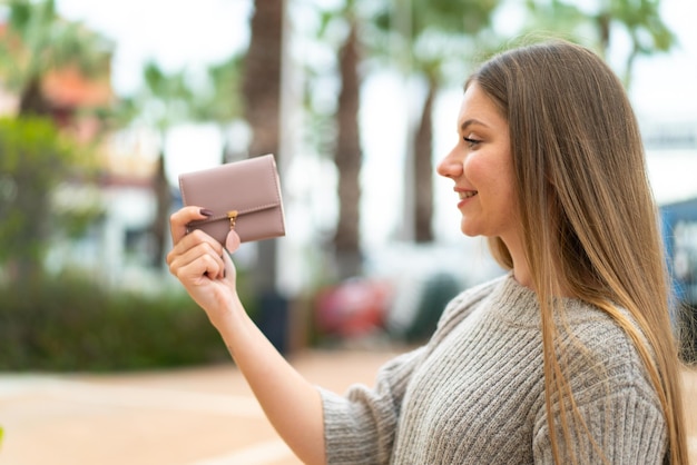 Junge hübsche blonde Frau, die draußen eine Brieftasche mit glücklichem Ausdruck hält