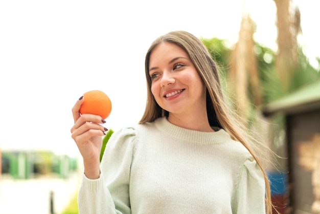 Junge hübsche blonde Frau, die draußen eine Orange mit glücklichem Ausdruck hält