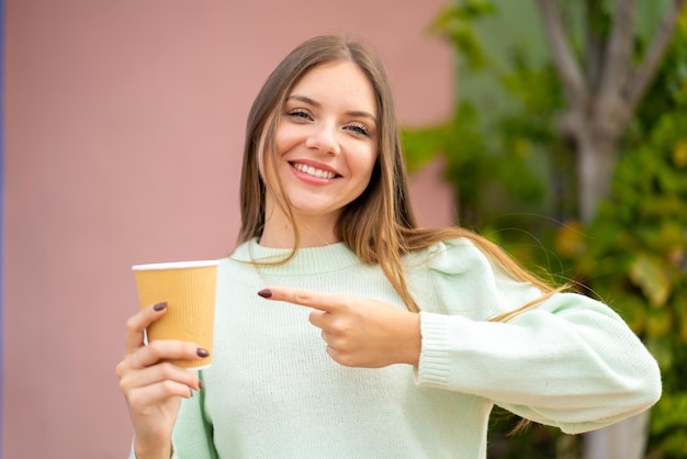 Junge hübsche blonde Frau, die draußen einen Kaffee zum Mitnehmen hält und darauf zeigt