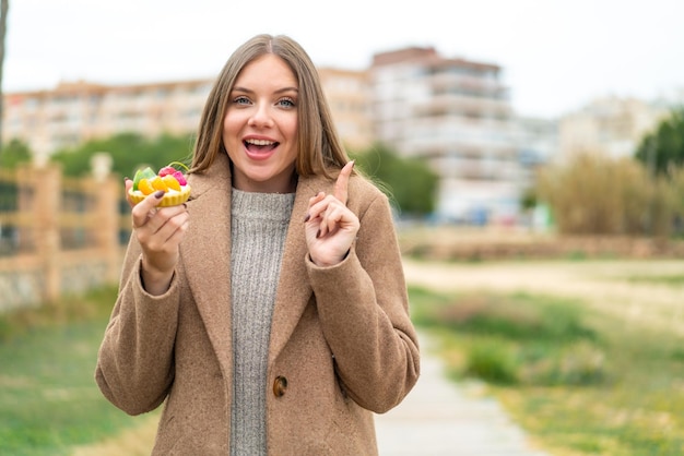 Junge hübsche blonde Frau, die ein Törtchen im Freien hält und auf eine großartige Idee zeigt