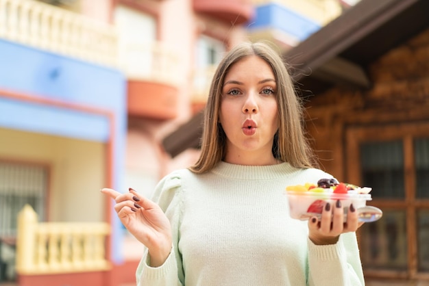 Junge hübsche blonde Frau, die eine Obstschale im Freien hält, überrascht und zeigt zur Seite