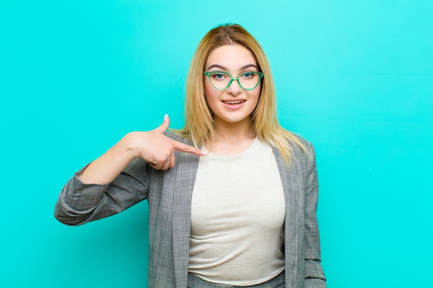 Junge hübsche blonde Frau, die glücklich, stolz und überrascht aussieht, fröhlich auf sich selbst zeigt, sich sicher und hoch gegen flache Wand fühlt