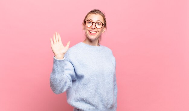 Junge hübsche blonde Frau, die glücklich und fröhlich lächelt, die Hand winkt, Sie begrüßt und begrüßt oder sich verabschiedet