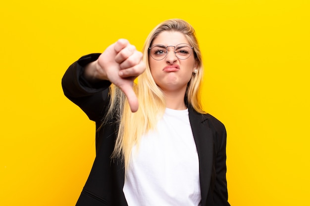 Junge hübsche blonde Frau, die sich böse, wütend, genervt, enttäuscht oder unzufrieden fühlt und Daumen nach unten mit einem ernsten Blick gegen die gelbe Wand zeigt