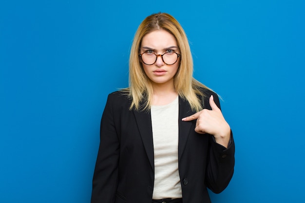 Junge hübsche blonde Frau, die sich verwirrt, verwirrt und unsicher fühlt, auf sich selbst zeigt und fragt, wer, ich? flache Wand